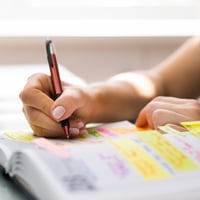 A book with words highlighted in yellow, pink, and purple is being written in with a brown pen.  